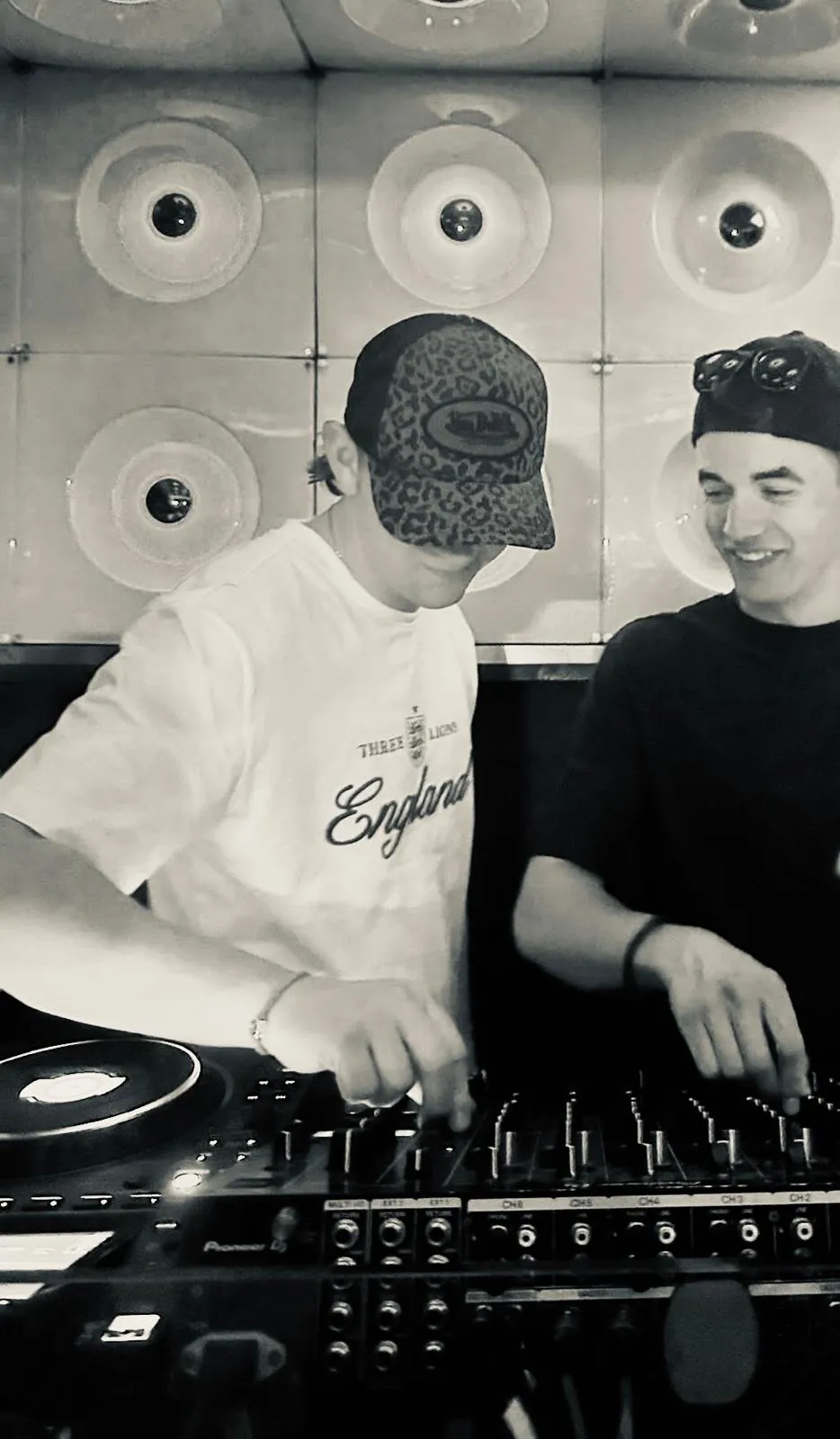 C@M playing back-to-back with another DJ on Pioneer decks, in front of a wall of mounted vinyl records