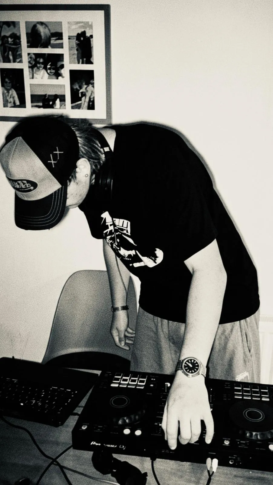 C@M in a cap and headphones, bent over a DJ controller at his home setup